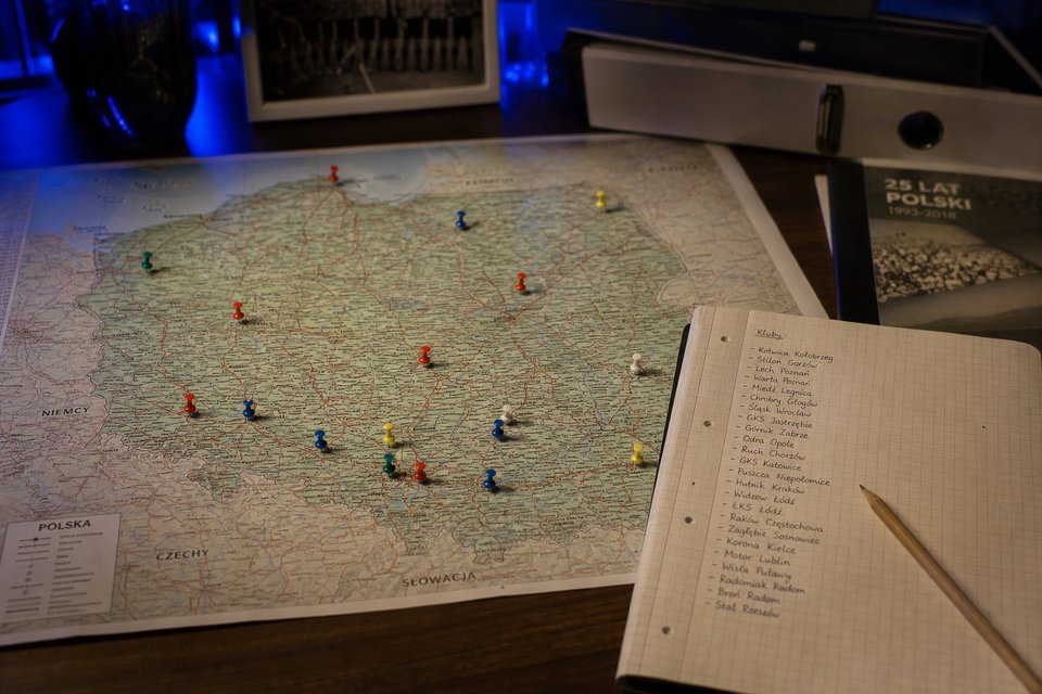 Papierowa mapa Polski z pinezkami obok notatnika z nazwami miejscowości, na biurku redakcyjnym